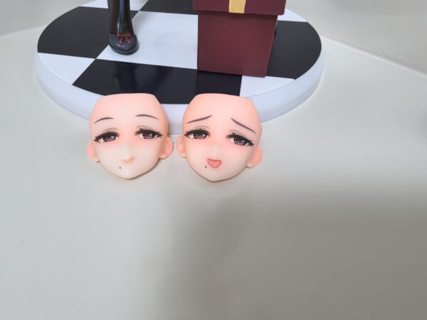 Two doll heads with detailed eyes and blush, displayed on a black-and-white checkered base